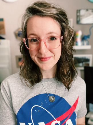 Libby is a white woman with wavy brown and grey hair and brown eyes. She's wearing a grey nasa shirt and pink glasses, and she's smiling warmly. A shelf behind her is full of colorful items.